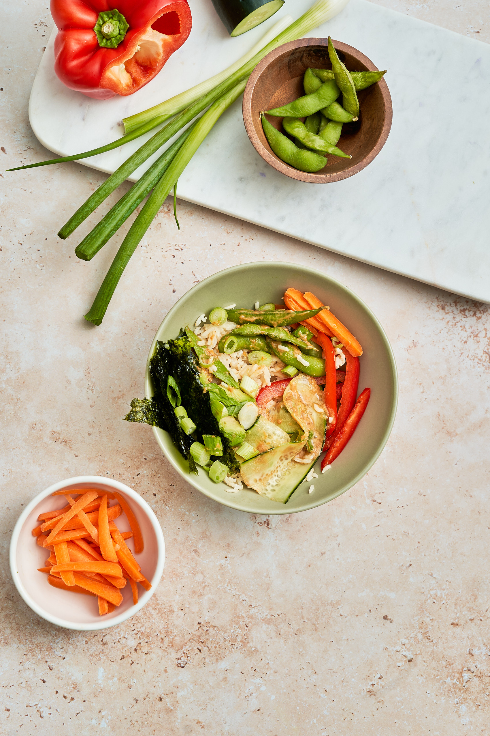 a cutting board with green onions on it next to a vegan sushi bowl and a small bowl of sliced carrots