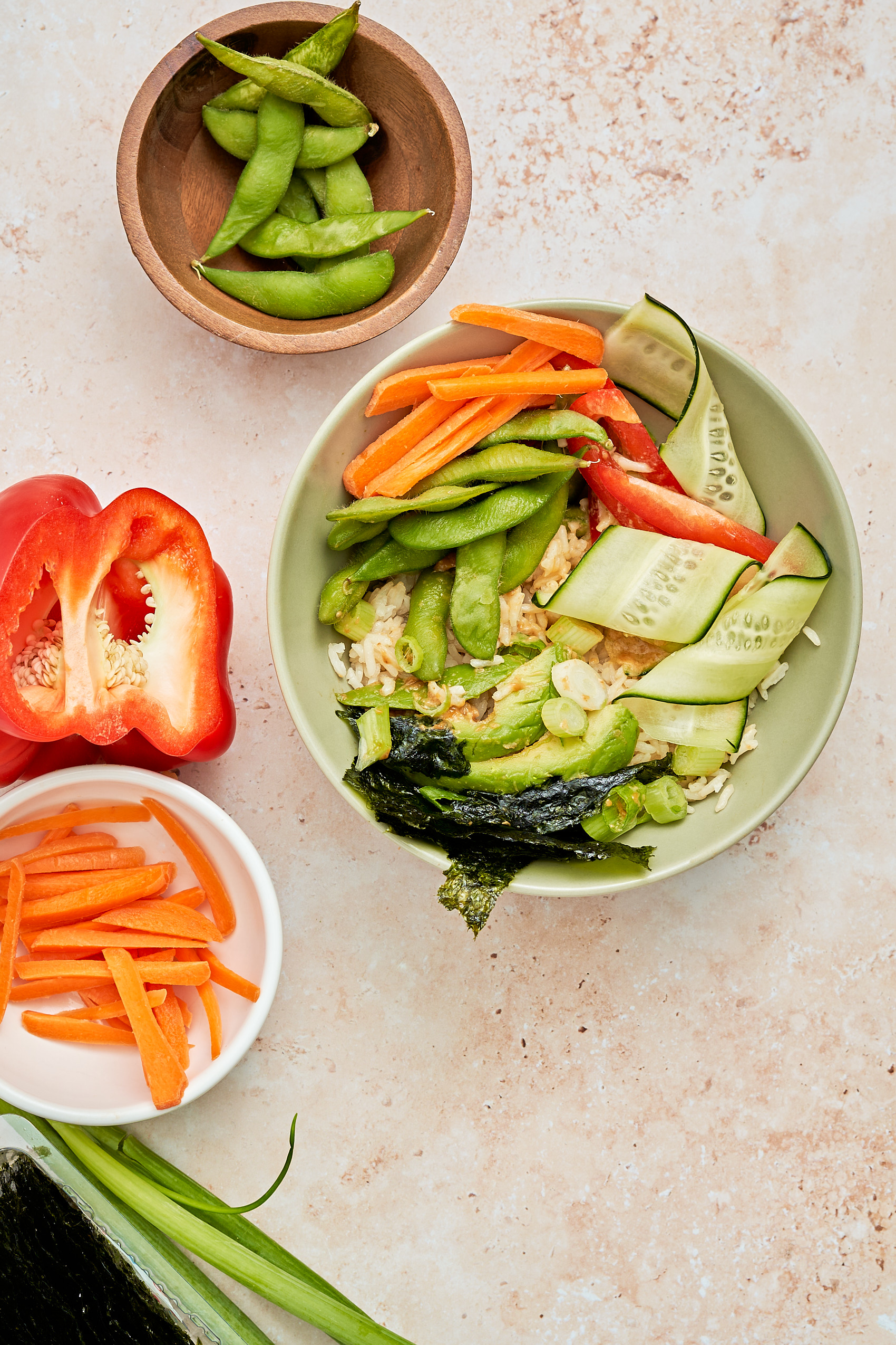 a vegan sushi bowl surrounded by a bell pepper, a bowl of sliced carrots and a bowl of edamame