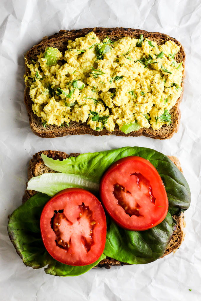 Two pieces of bread about to be stacked into a sandwich, one half topped with lettuce and tomatoes and the other half topped with vegan egg salad