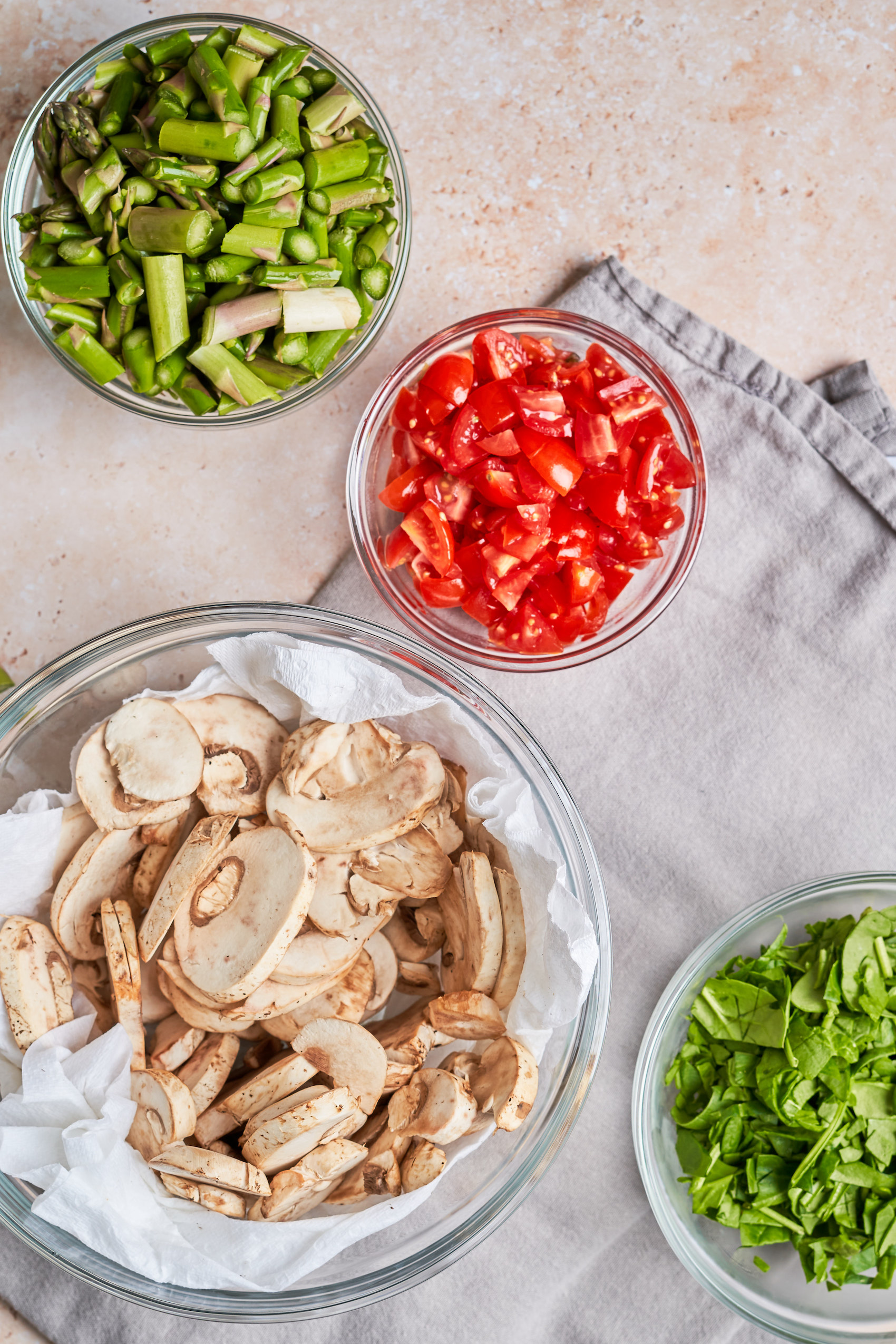 a collection of bowls holding raw vegetables including sliced mushrooms, asparagus, tomatoes and greens