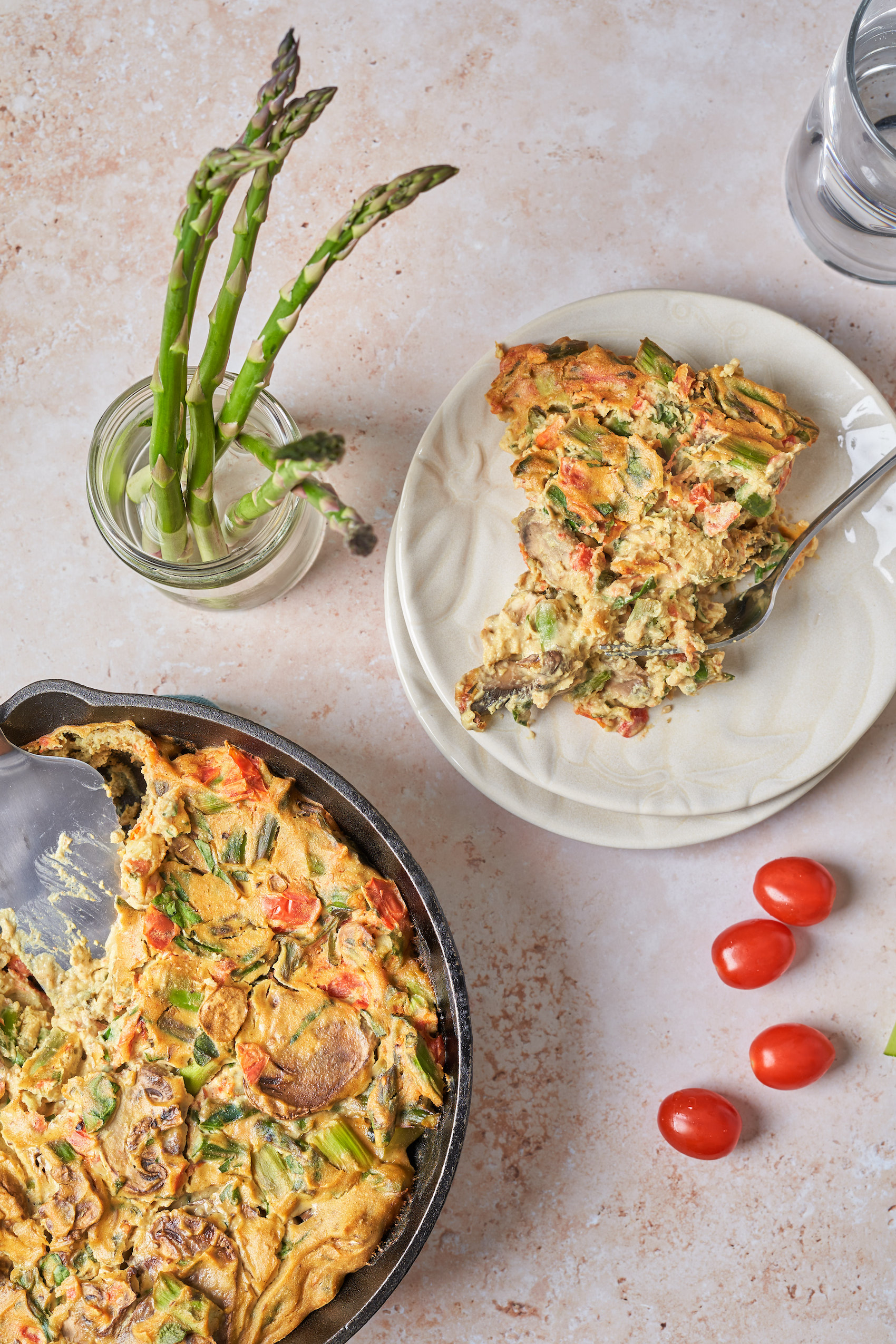 a slice of vegan quiche on a plate next to a cast iron skillet holding the rest of the quiche alongside a few cherry tomatoes and a glass jar of asparagus