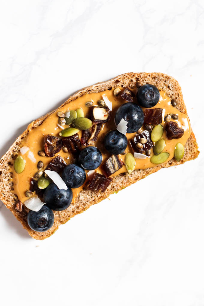 A piece of whole grain toast topped with peanut butter, chopped dates, blueberries and seeds