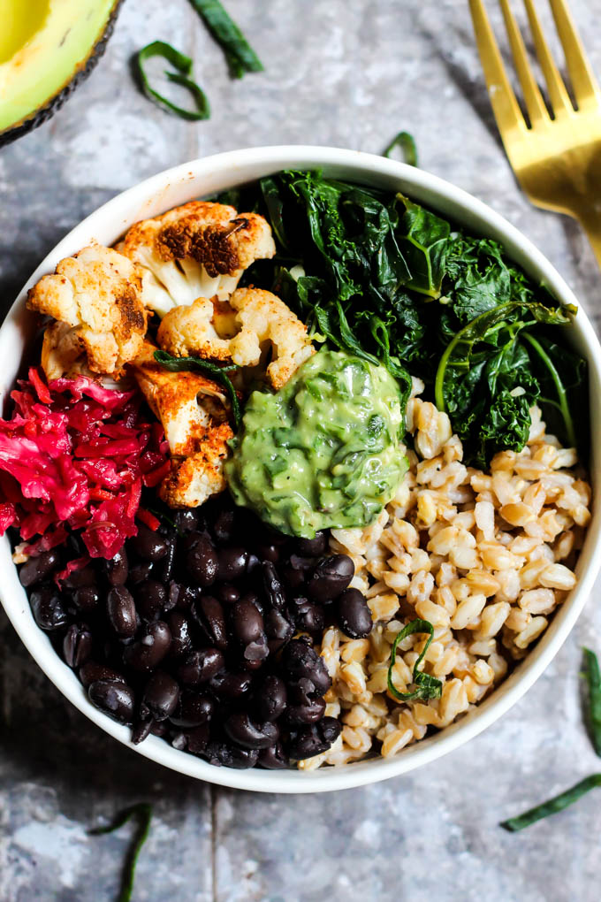 A close up shot of a power bowl made with roasted cauliflower, black beans, cooked kale, farro, sauerkraut and a dollop of homemade avocado pesto