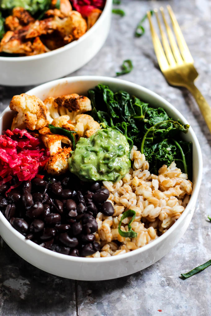 A close up of a power bowl made with black beans, farro, wilted kale, cauliflower and sauerkraut served with a dollop of avocado pesto