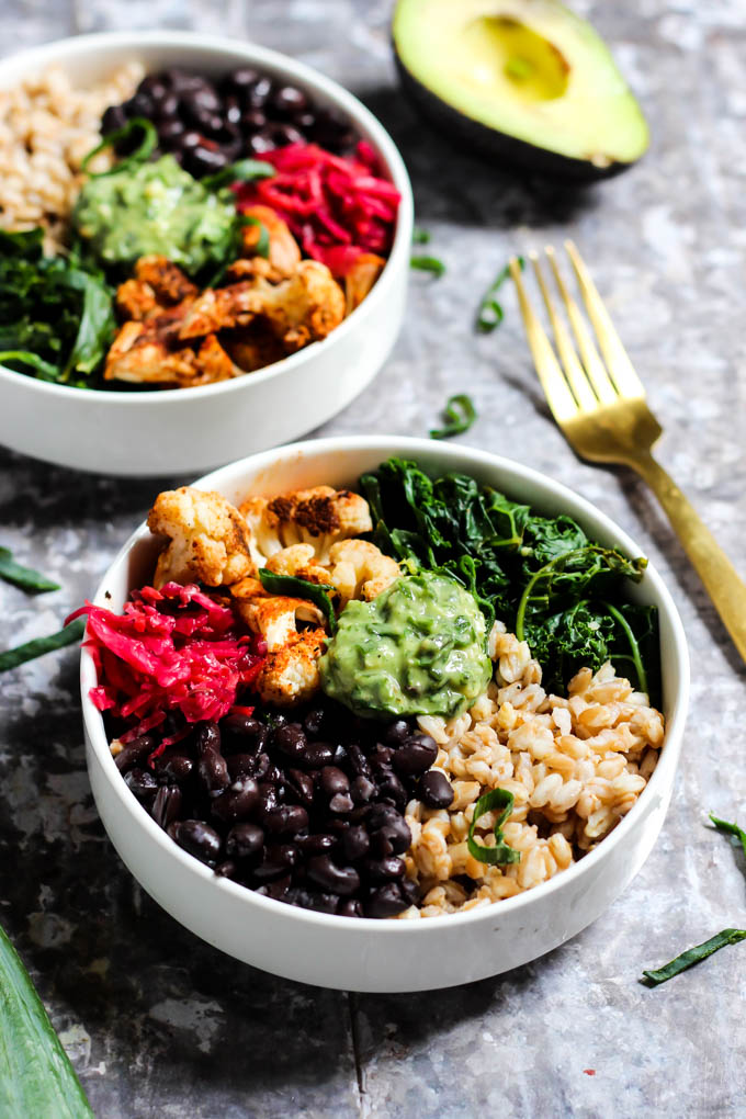 Two power bowls containing farro, black beans, sauerkraut, roasted cauliflower and wilted kale topped off with a dollop of avocado pesto