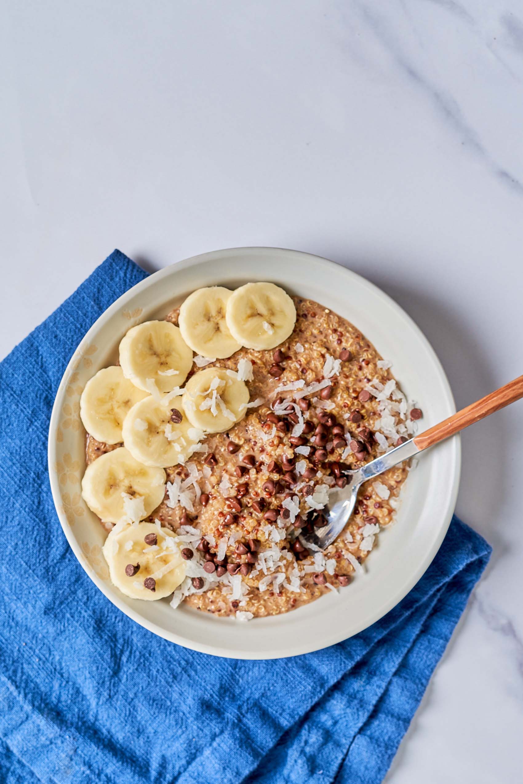 a spoon in a bowl of quinoa porridge topped with bananas, chocolate chips and coconut