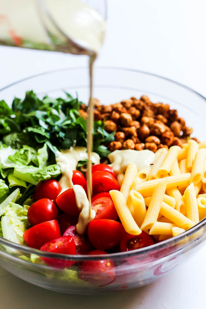 A large bowl of romaine, tomatoes, parsley, roasted chickpeas and penne pasta being drizzled with a dairy free caesar dressing