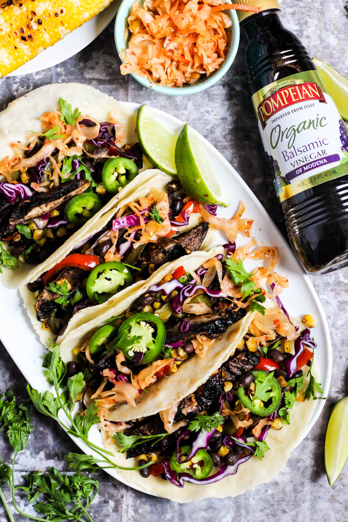 a platter of mushroom tacos served with cilantro, kimchi, corn and limes alongside a bottle of balsamic vinegar