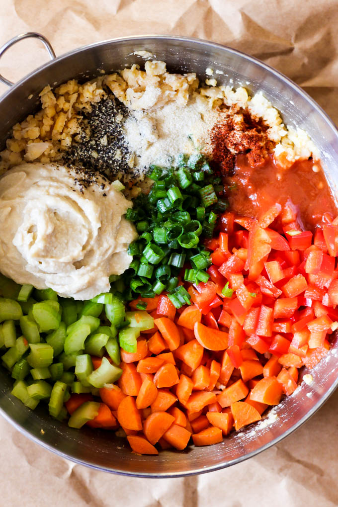 A large mixing bowl filled with hummus, spices, chopped carrots, peppers, green onion, celery, hot sauce and chickpeas