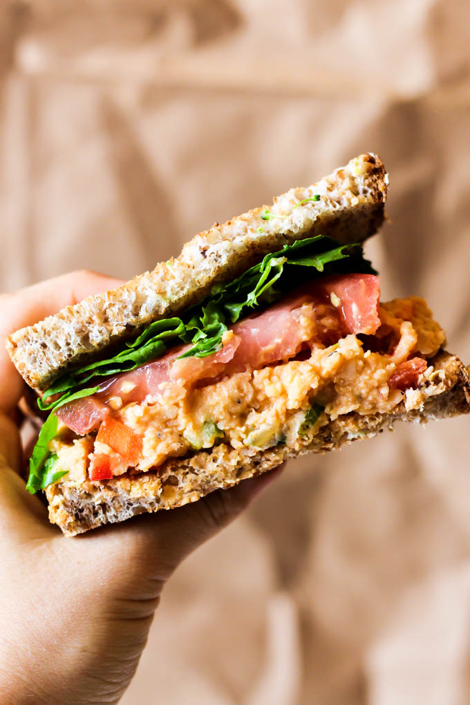 a hand holding a vegan sandwich topped with mashed chickpeas, lettuce and tomatoes