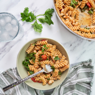 a bowl of pasta served with tomatoes, kale and white beans next to a larger skillet full of pasta