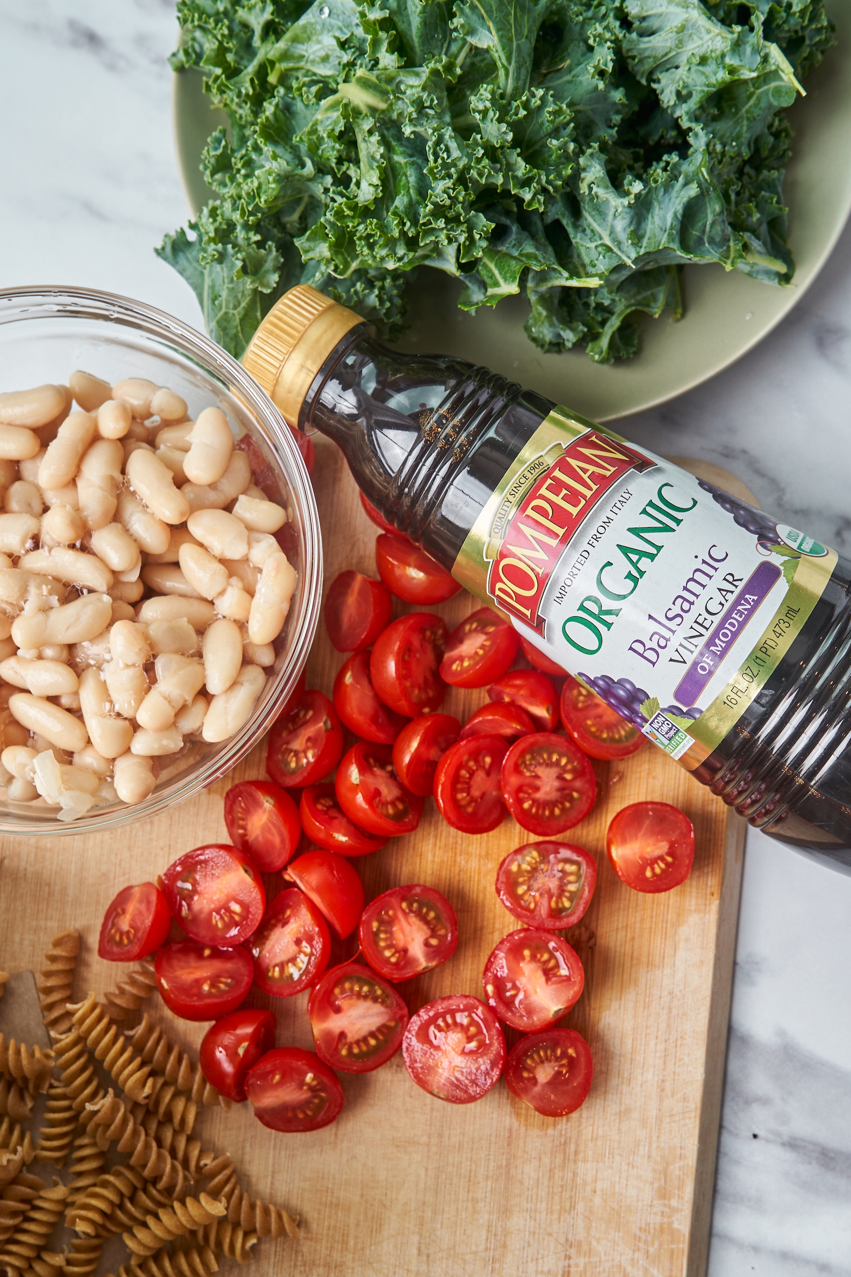 a cutting board filled with halved cherry tomatoes, a bottle of vinegar, a bowl of drained white beans and a bundle of kale