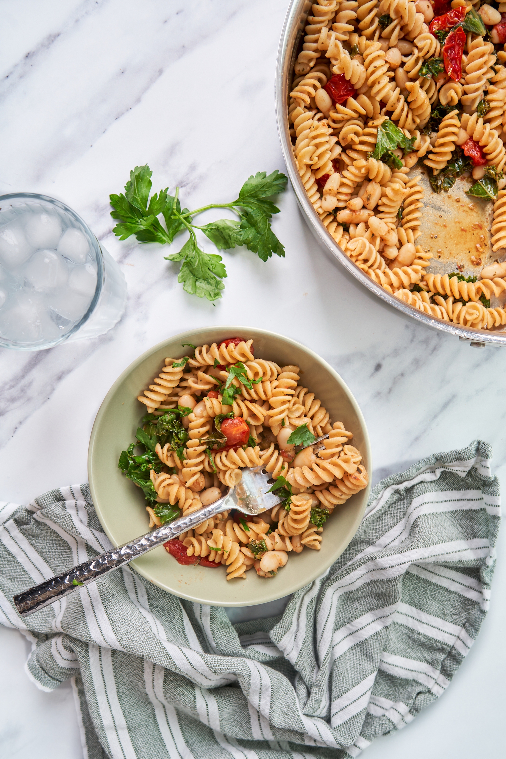 a bowl of fusilli pasta served with tomatoes, kale and white beans next to a larger skillet filled with pasta and a sprig of fresh parsley