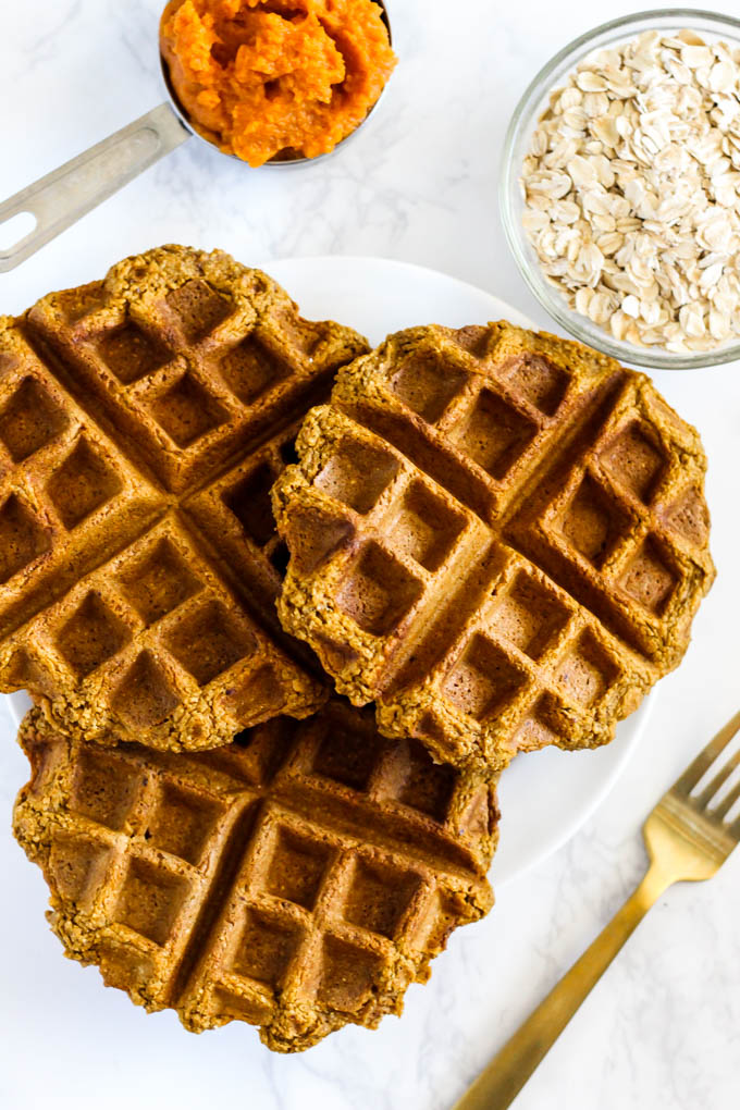 three pumpkin waffles on a plate next to a bowl of oats and a measuring cup of pumpkin