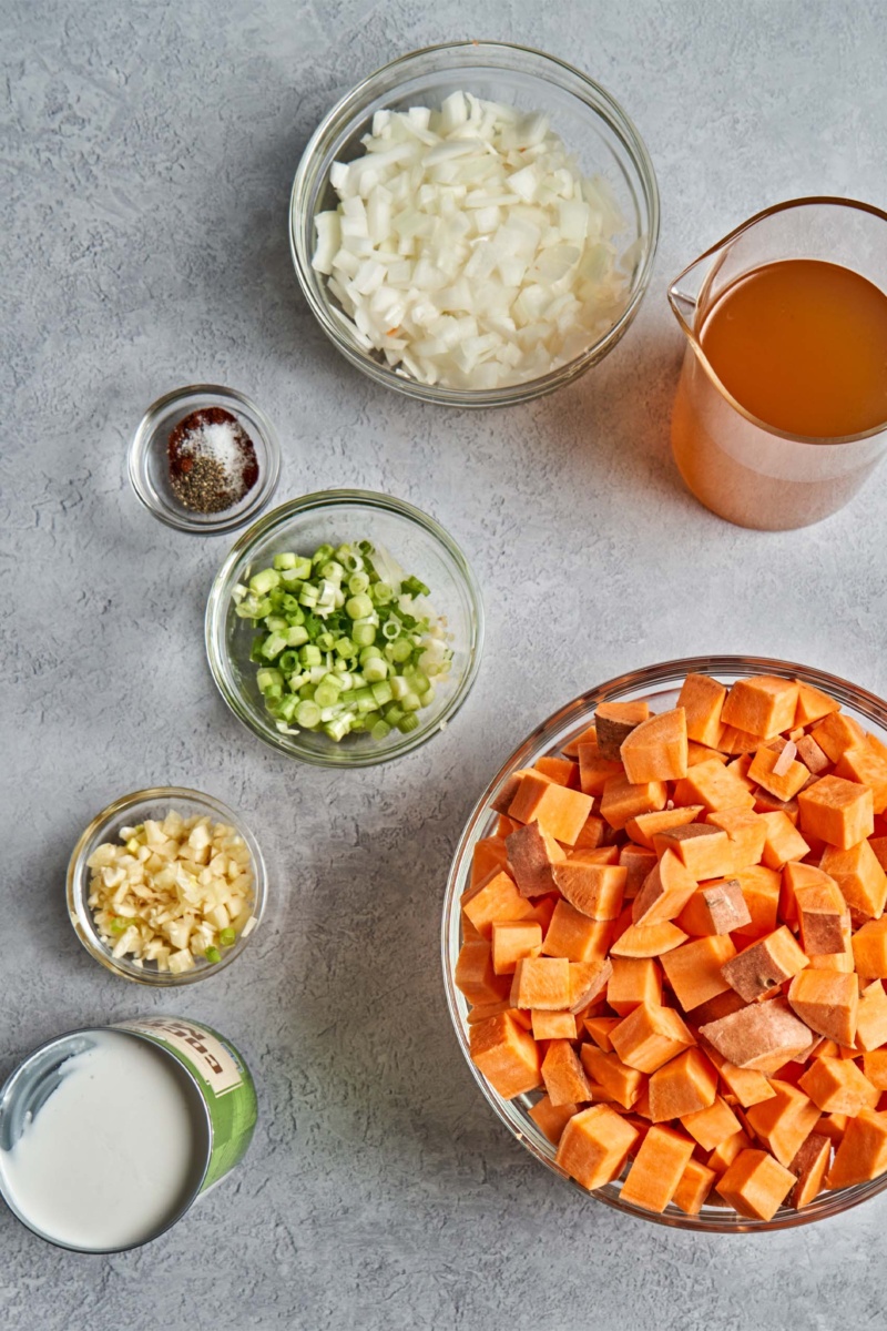 a bowl of sweet potatoes, a small bowl of chopped garlic, a bowl of sliced green onions, a bowl of chopped onions, a cup of coconut milk, a jug of vegetable broth and a small dish of spices