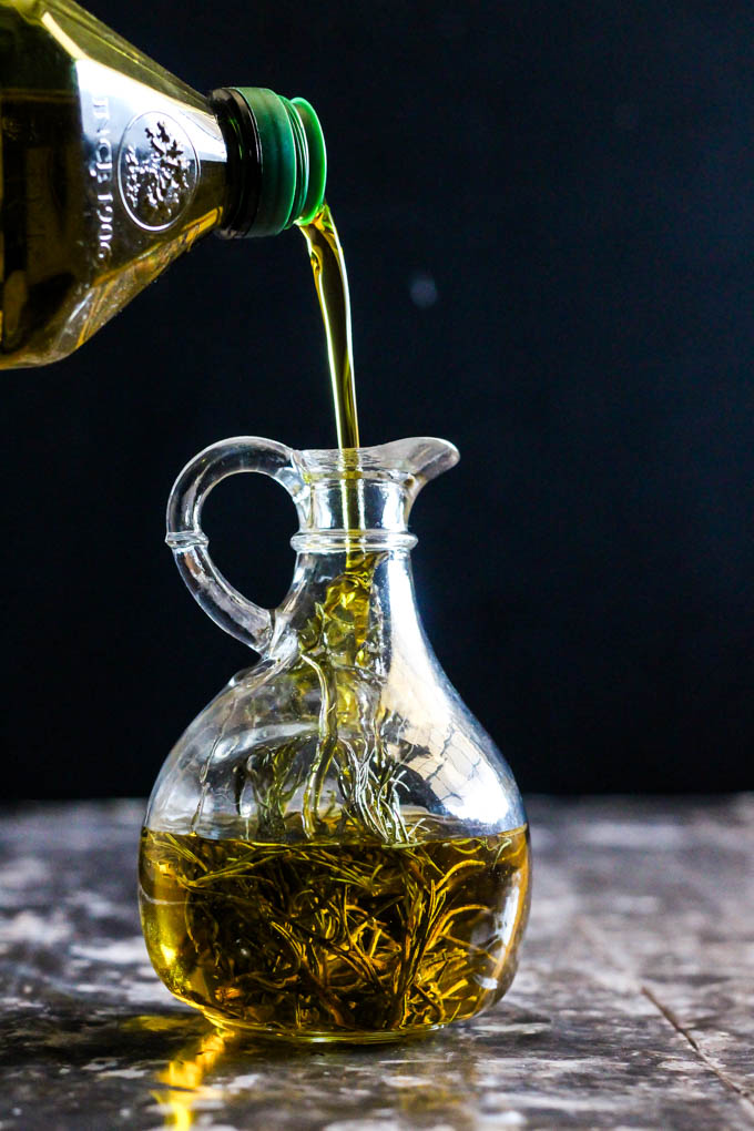 a bottle of olive oil being poured into a class bottle containing rosemary