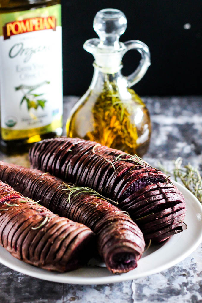a plate of hasselback sweet potatoes garnished with rosemary nerved next to a bottle of rosemary infused olive oil