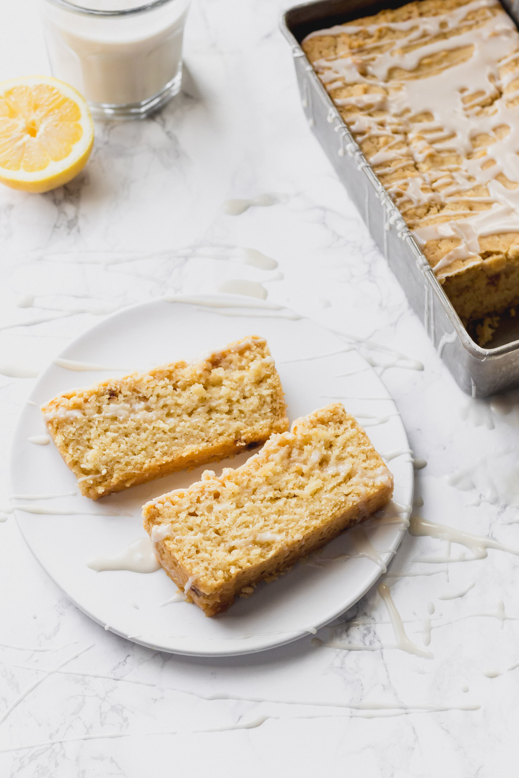 two servings of lemon cake resting on a plate next to the tin holding the full loaf