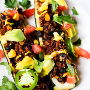 two halved zucchinis filled with a Southwestern mixture of vegan ground meat, beans, corn, avocado, tomatoes and a drizzle of vegan nacho cheese