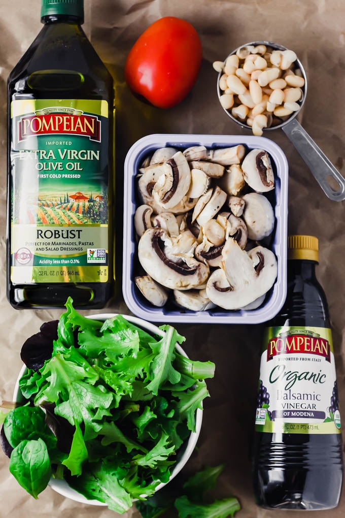 a measuring cup full of white beans, a tomato, a carton of mushrooms, a bowl of greens and bottles of balsamic vinegar and olive oil