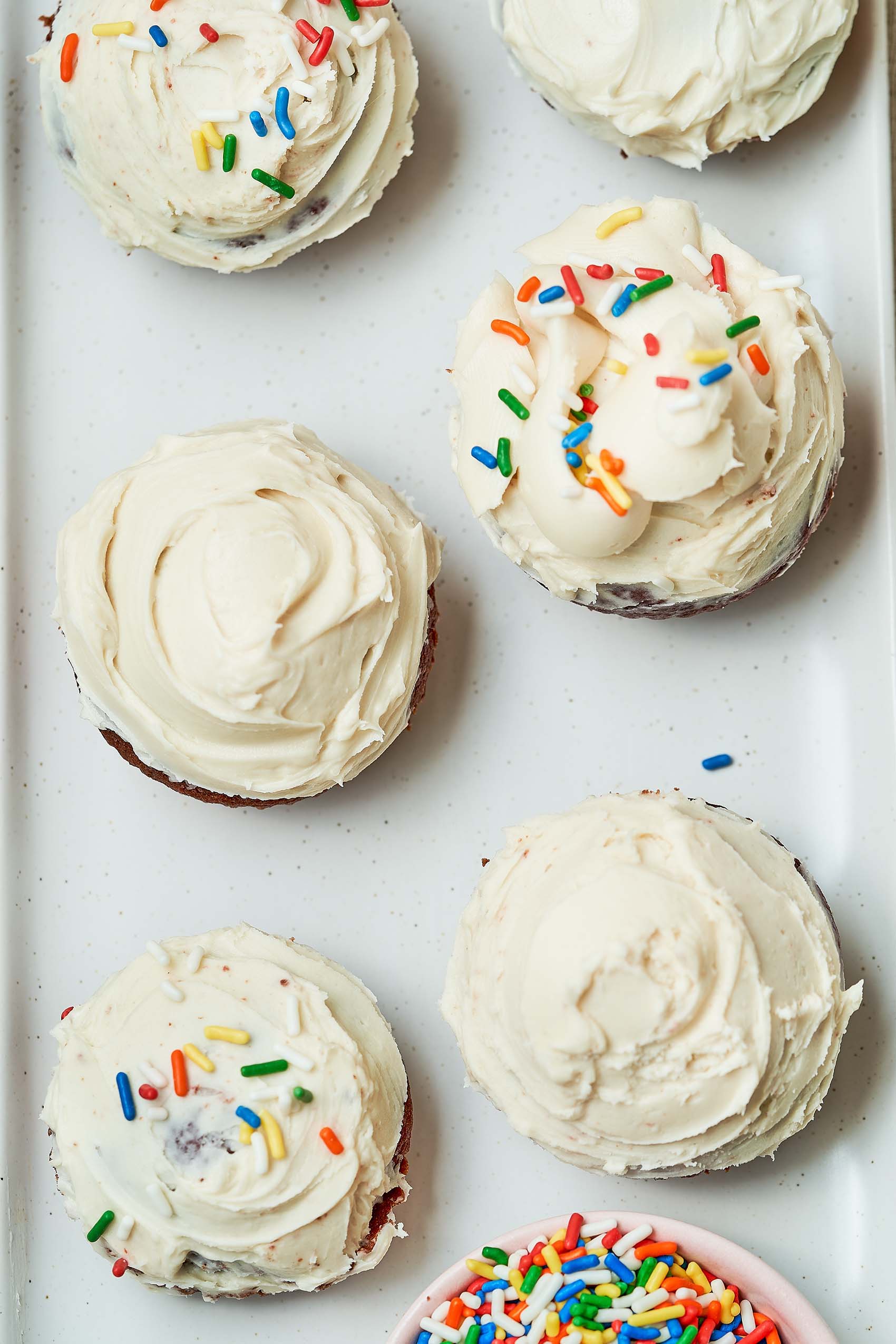 an overhead shot of a batch of cupcakes topped with vanilla icing and sprinkles resting on a white tray