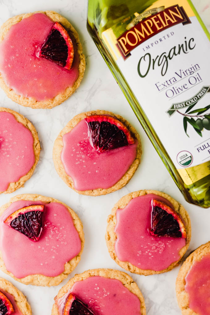 sugar cookies topped with pink glaze and a blood orange segment laid next to a bottle of olive oil