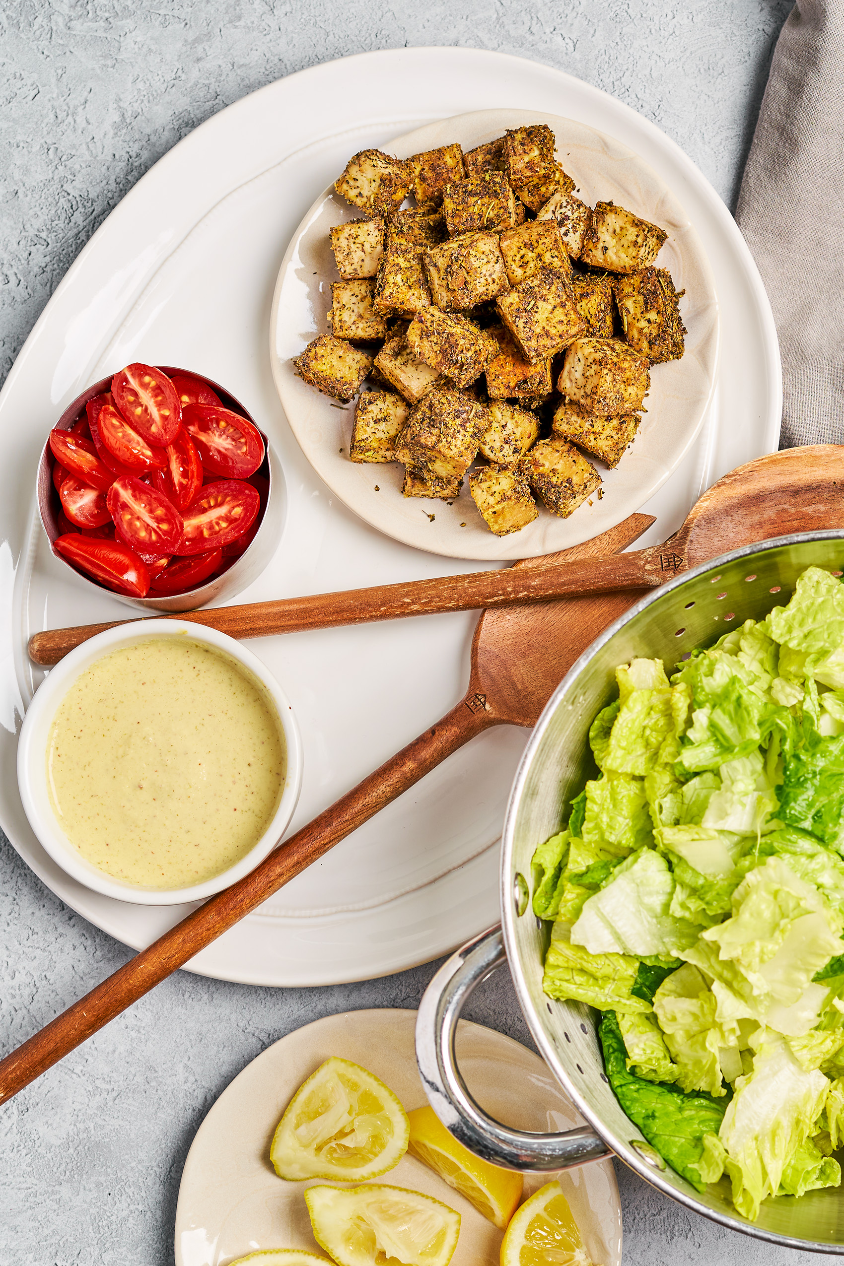 a serving platter of crispy baked tofu, sliced cherry tomatoes and caesar dressing next to a large bowl of romaine