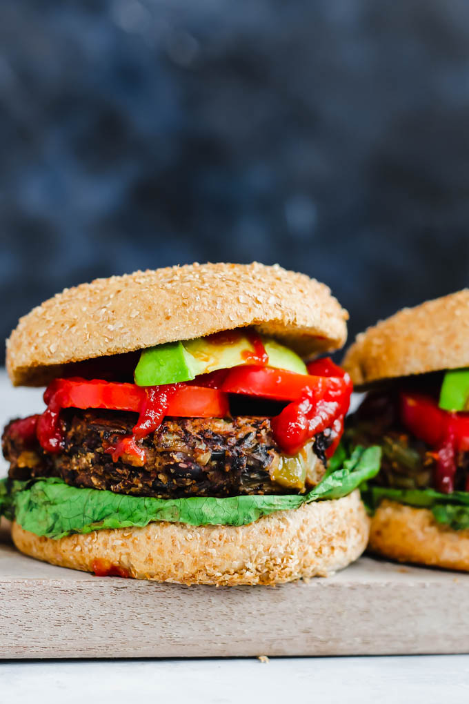 Two mushroom wild rice veggie burgers topped with a slice of avocado, two slices of tomato, a drizzle of ketchup and lettuce served on a wooden cutting board