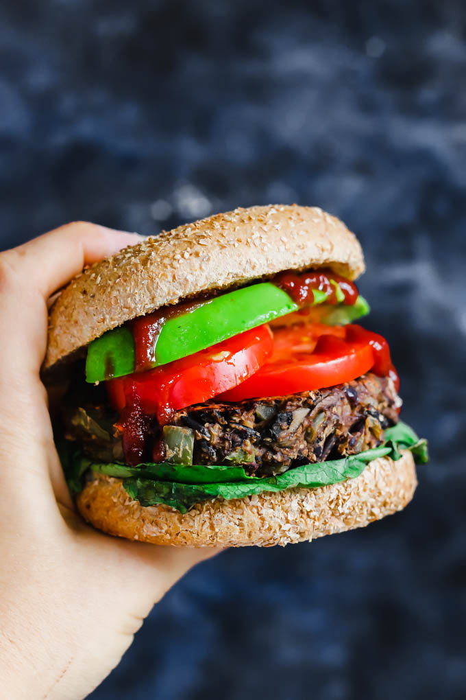 A hand holding a wild rice mushroom veggie burger topped with ketchup, avocado, tomato and lettuce