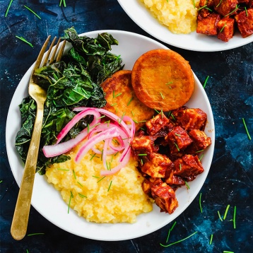 a bowl of grits, tofu, collard greens, sweet potato slices and pickled onions with a fork