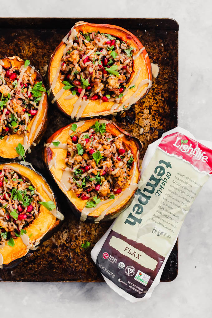 a tray of halved acorn squashes, each filled with a tempeh and wild rice stuffing and topped with cilantro and a drizzle of tahini