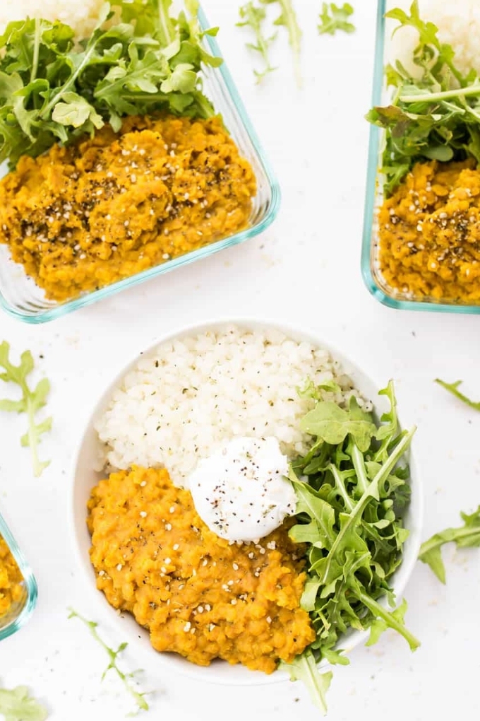 Vegan red lentil dal is served in a white bowl and two meal prep containers alongside white rice and arugula