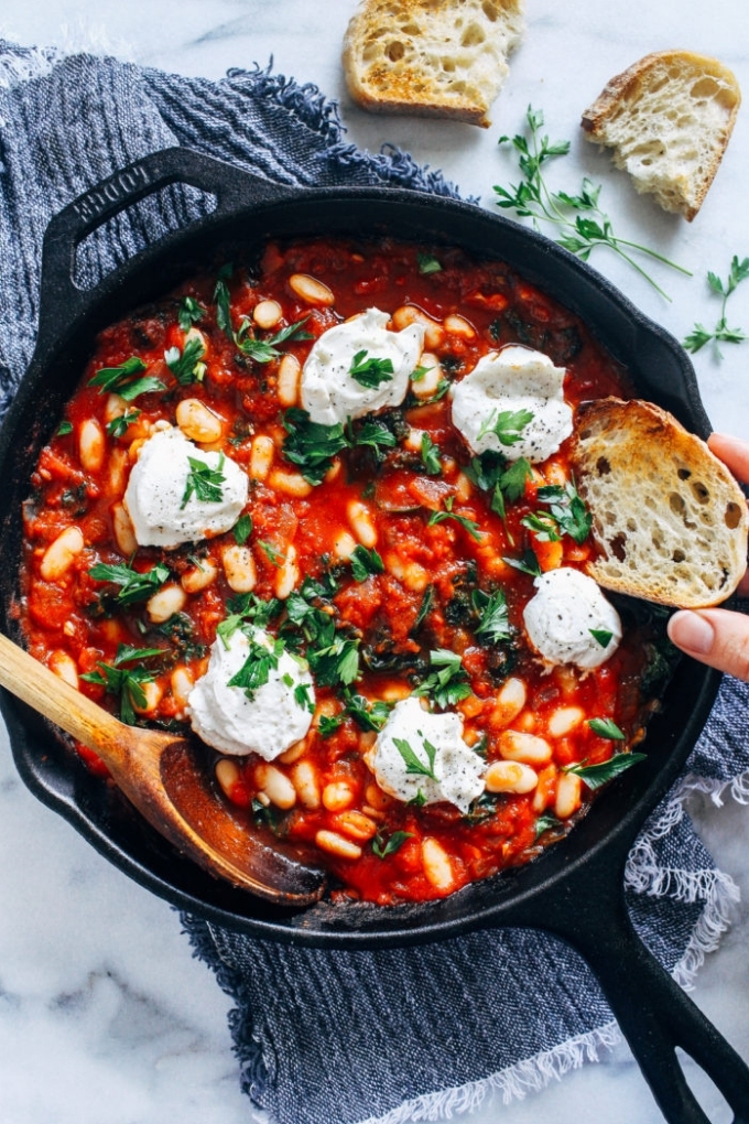 A hand holding a slice of bread scoops into a cast iron skillet of vegan shakshuka