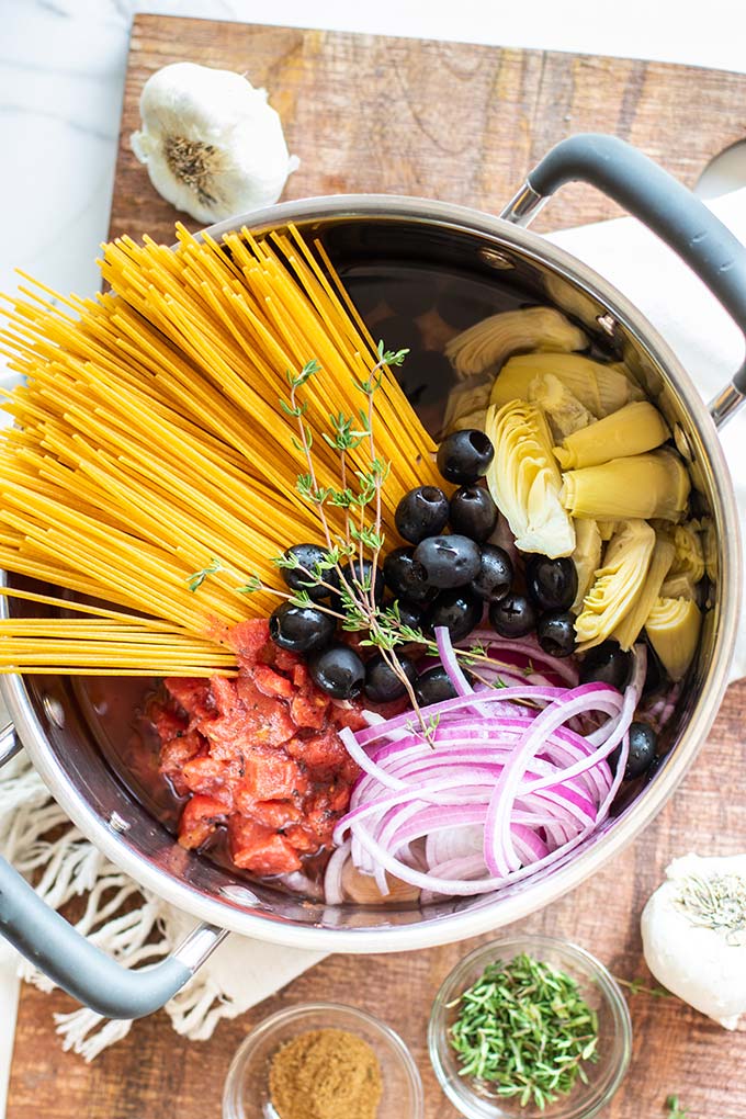 A pot filled with dried spaghetti, red onion, black olives, artichokes, tomatoes and sprigs of thyme sits on a cutting board surrounded by bulbs of garlic, thyme and spices