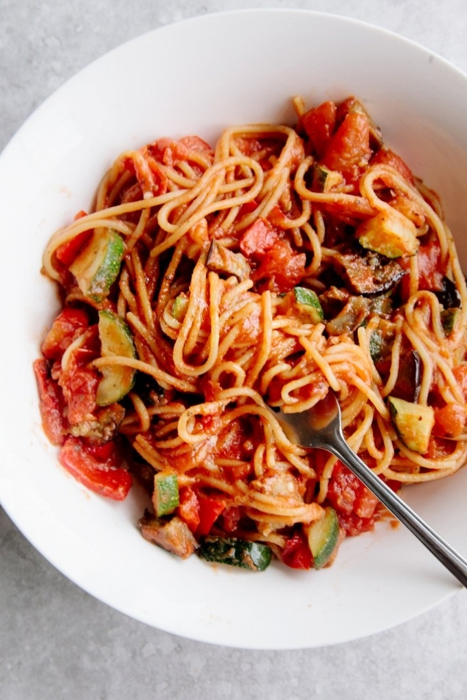 A fork digging into a white bowl filled with spaghetti, tomato sauce and veggies