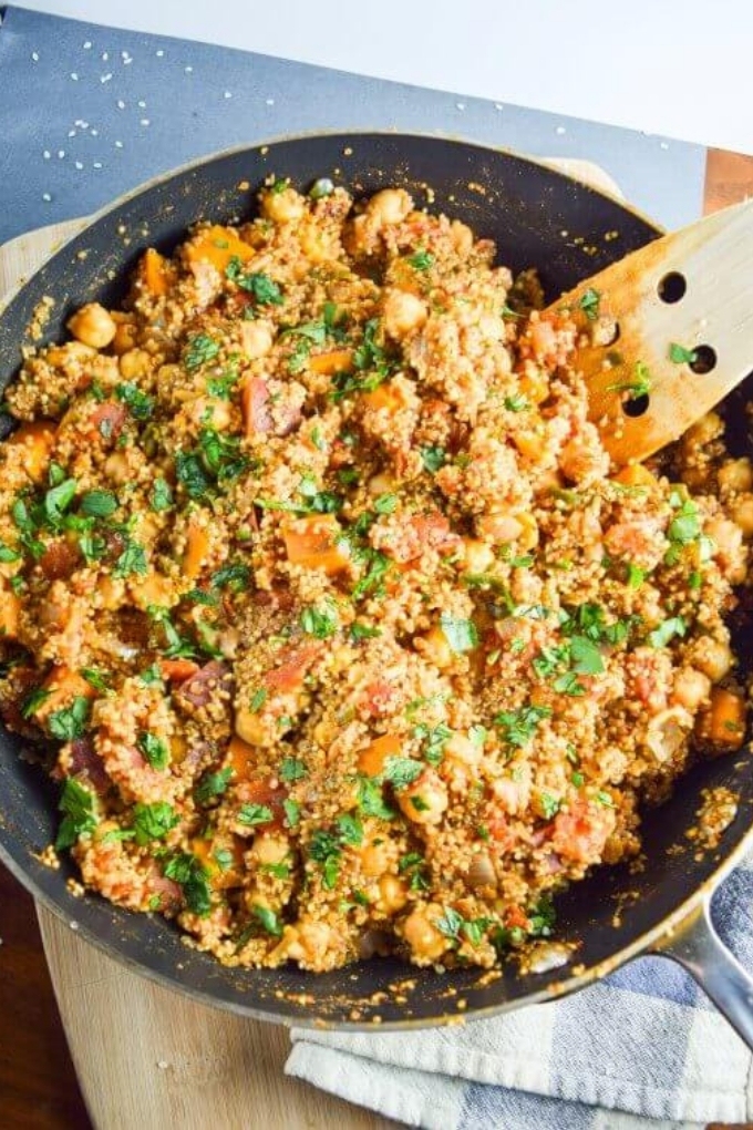 A skillet filled with cooked quinoa, tomatoes and herbs being stirred with a wooden spatula