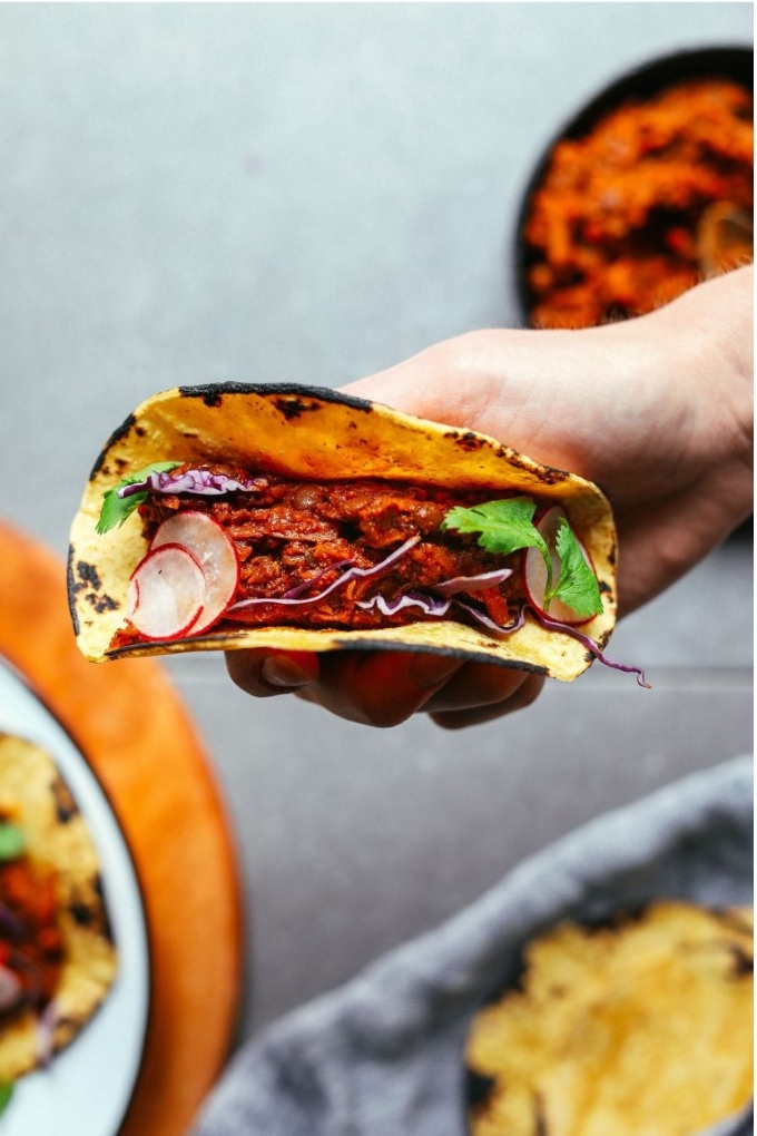 A hand holding a corn tortilla filled with vegan barbacoa, sliced radishes and cilantro