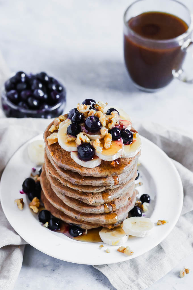 Sliced bananas, blueberries and walnuts top off a stack of buckwheat pancakes served alongside a cup of coffee and a bowl of blueberries