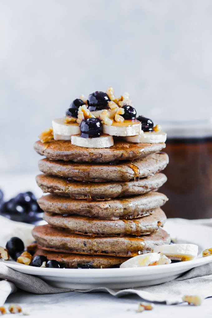 A stack of seven buckwheat pancakes is served with fruit, nuts and a drizzle of maple syrup
