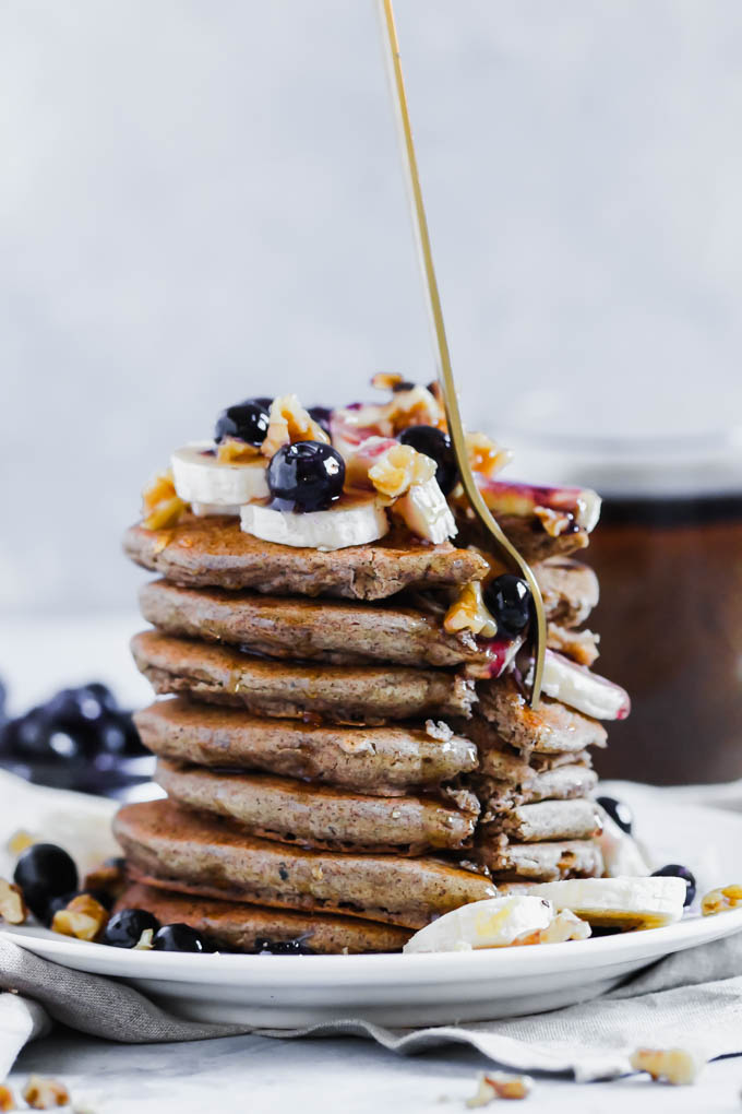 A fork digs into a stack of pancakes topped with fruit, nuts, and syrup