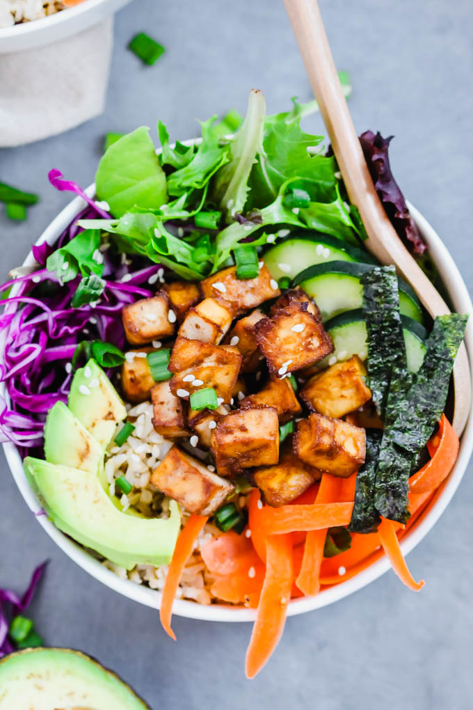 a bowl filled with roasted tofu, carrots, avocado slices, cabbage, greens, cucumber and nori