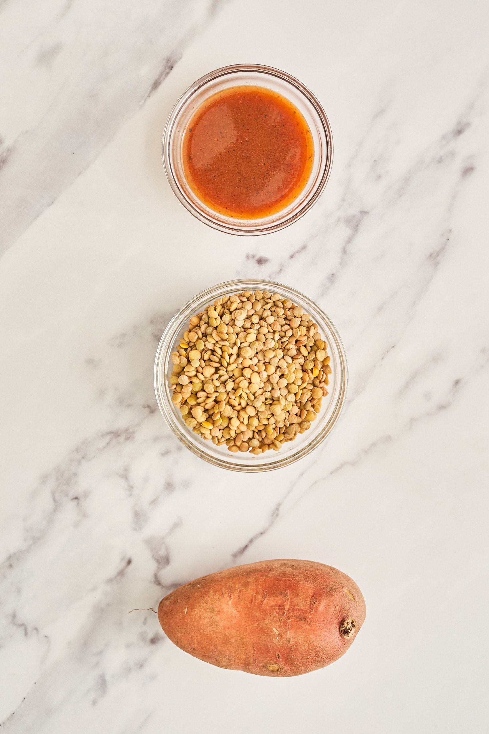 a selection of ingredients separated into small bowls, including a bowl of hot sauce, a bowl of green lentils and a whole sweet potato