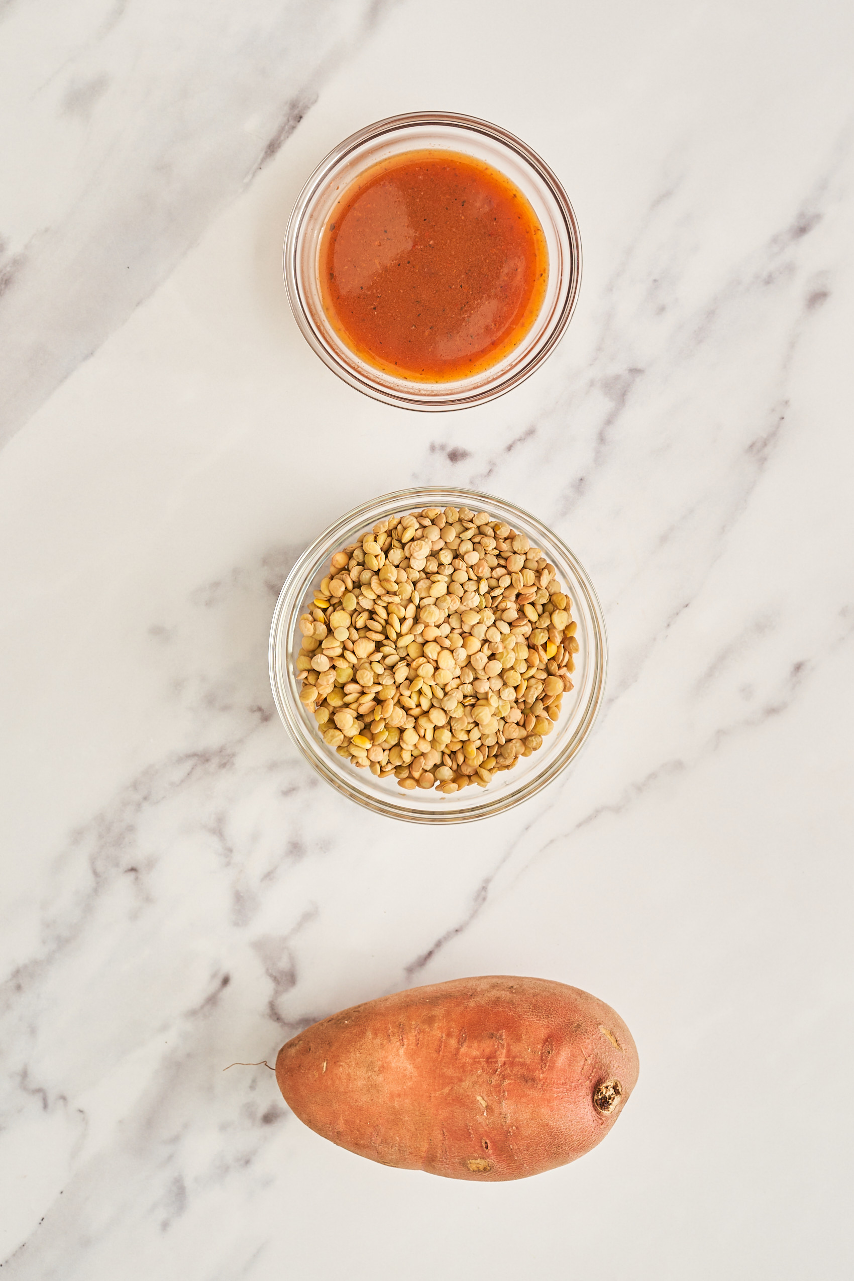 a selection of ingredients separated into small bowls, including a bowl of hot sauce, a bowl of green lentils and a whole sweet potato