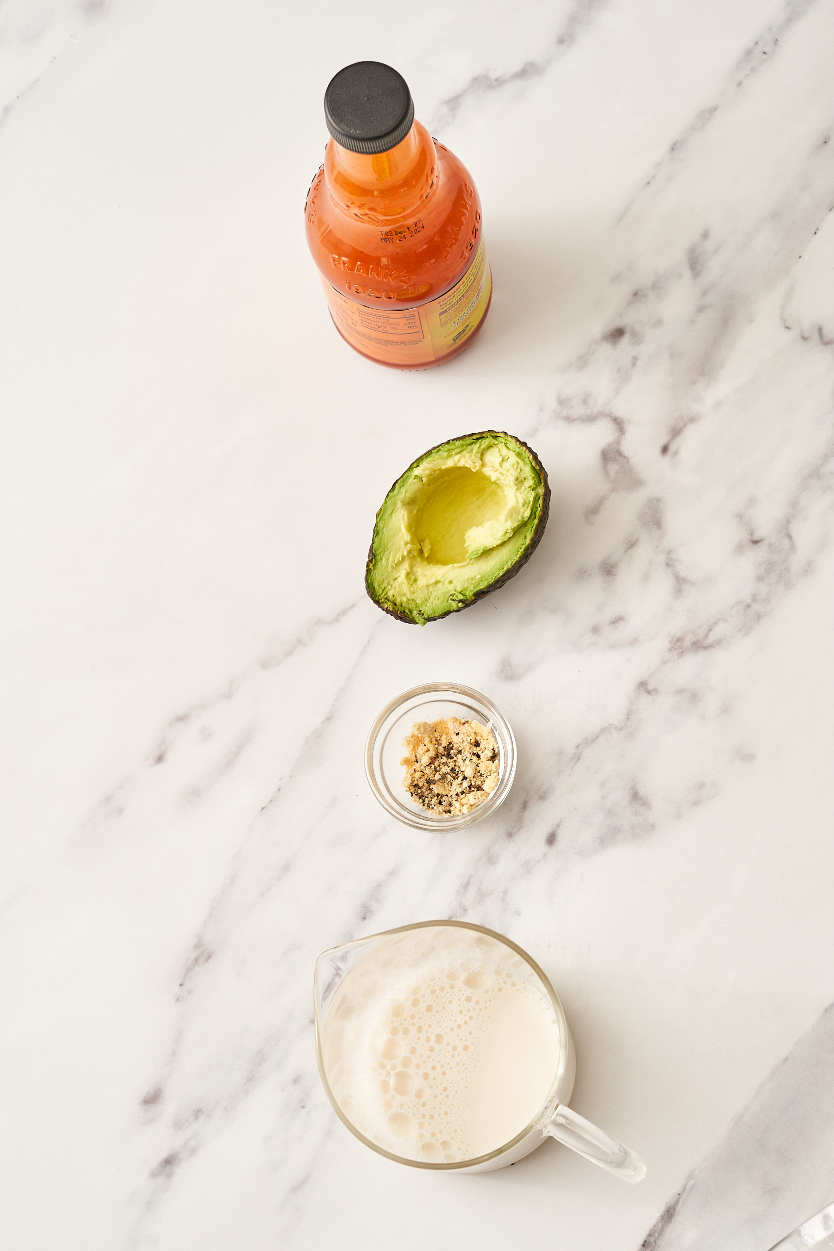 a bottle of hot sauce, a half an avocado, a small bowl of seasonings, and a large glass measuring cup of plant-based milk