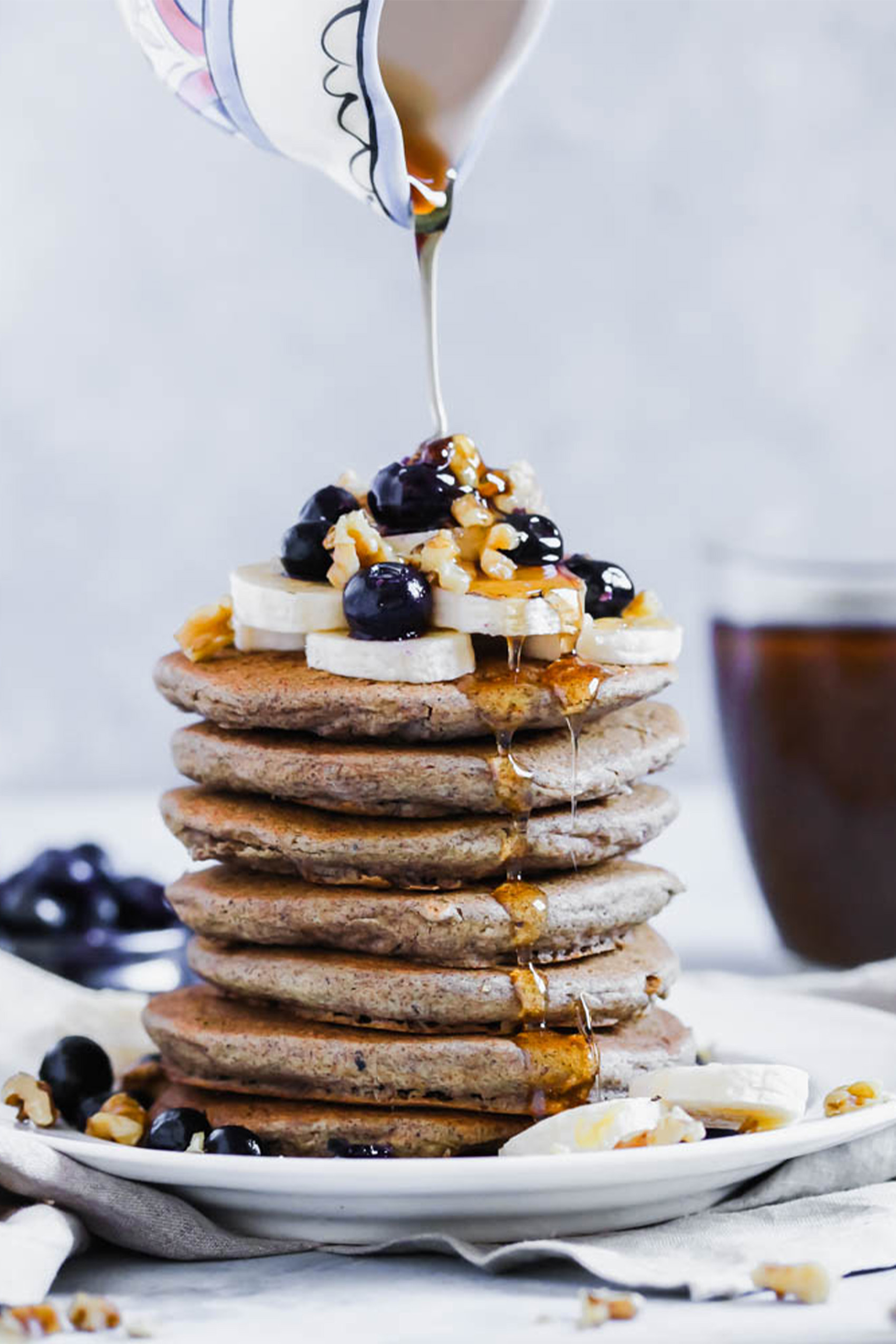 a stack of buckwheat pancakes topped with bananas, blueberries, walnuts and a drizzle of maple syrup