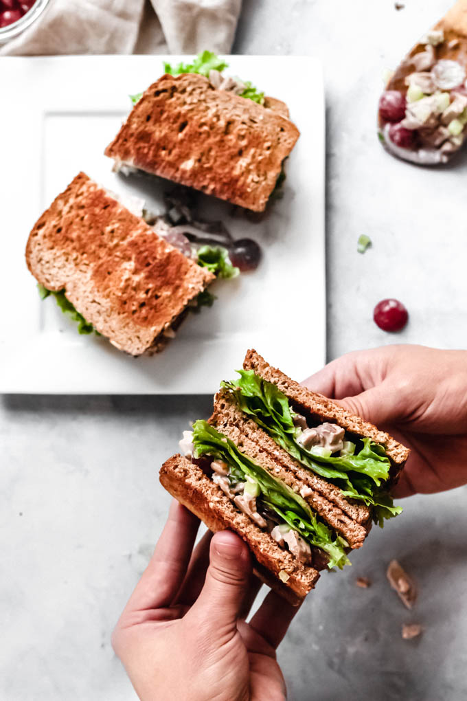 a hand holding a vegan a chicken salad sandwich next to a plate with a second sandwich served on it