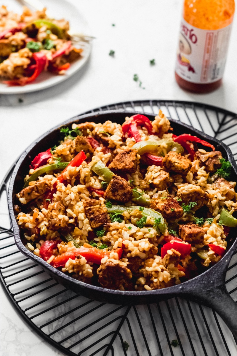 A cajun tempeh and rice skillet is served alongside a bottle of hot sauce