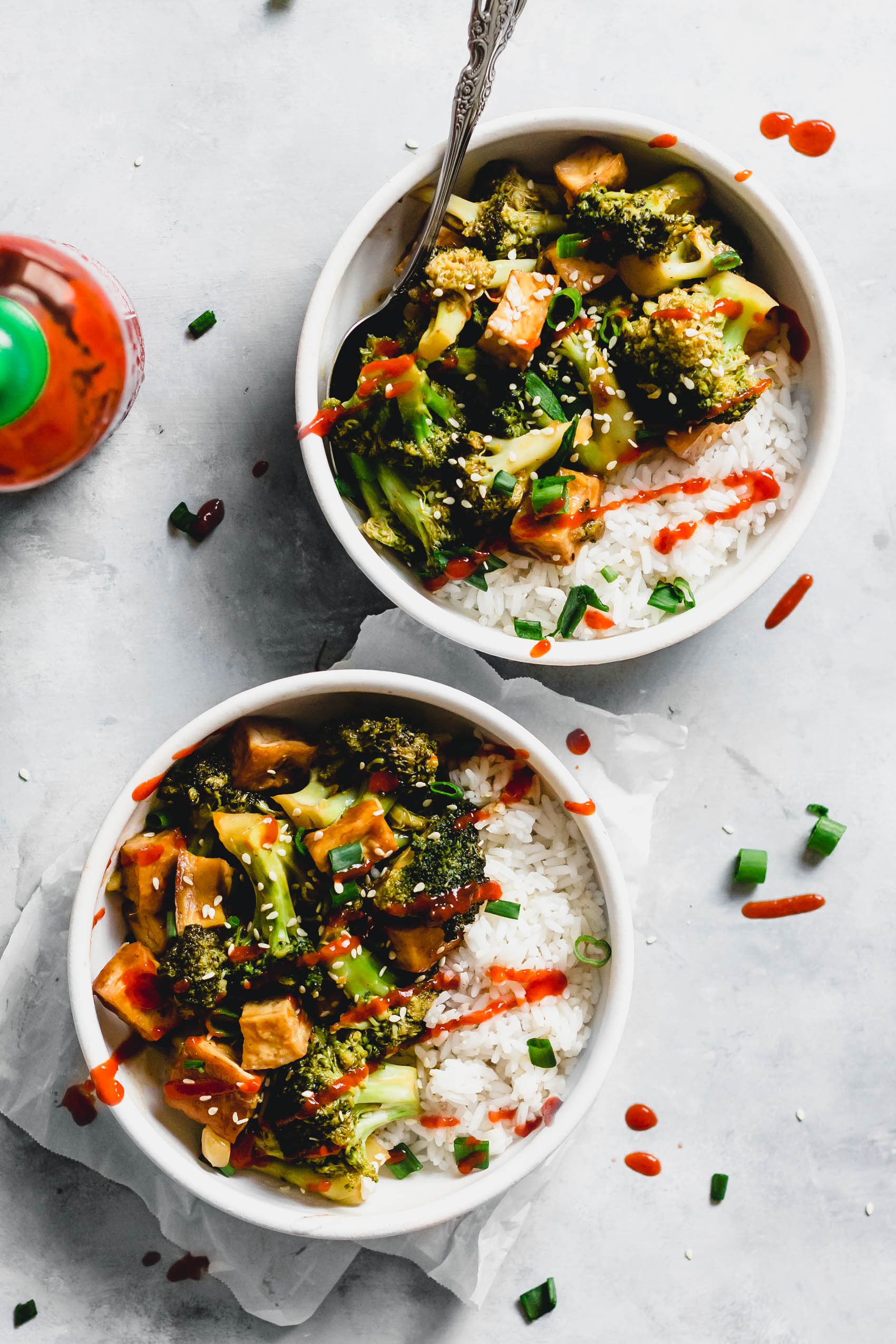 two servings of broccoli tofu stir fry served with sriracha and sliced green onion