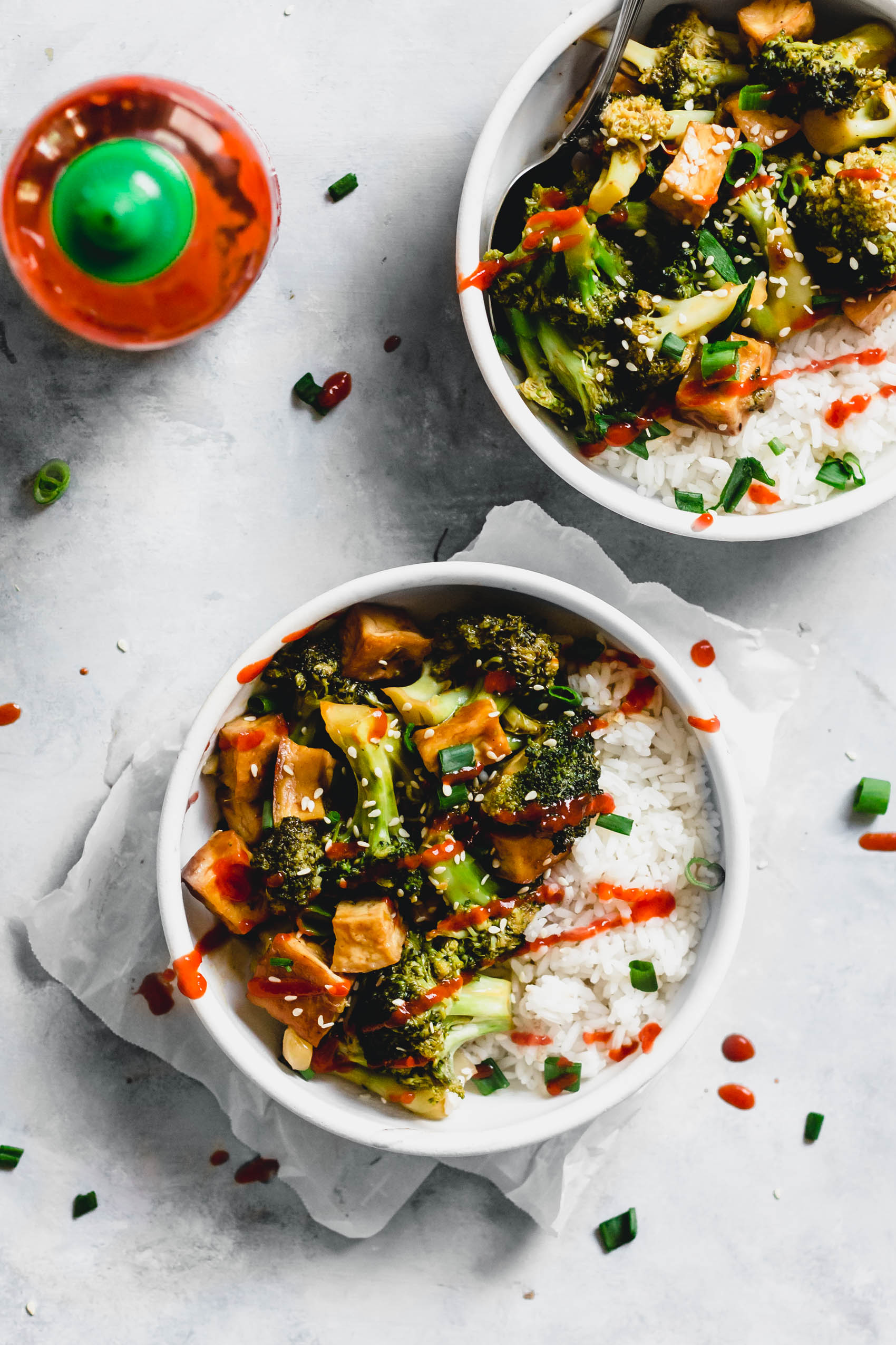 two bowls of tofu and broccoli stir fry served with green onion and sriracha