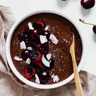 a wooden spoon resting in a bowl of chocolate chia pudding that is topped with halved cherries and coconut flakes